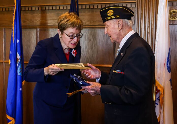 A woman (left) handing a man a piece of paper. The man is holding a folded American Flag with a suit on.